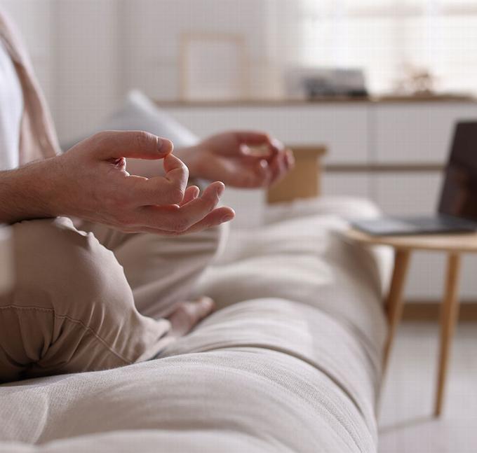 Person meditating on a sofa with hands in a calm gesture, laptop nearby.