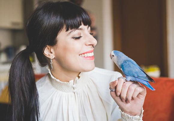 Woman smiling and interacting with a blue parakeet on her hand.