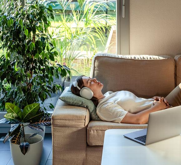 Relaxed person with headphones lying on a sofa next to a laptop and indoor plants.