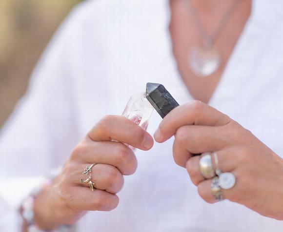 Two hands holding a clear quartz crystal and a black crystal, adorned with rings.