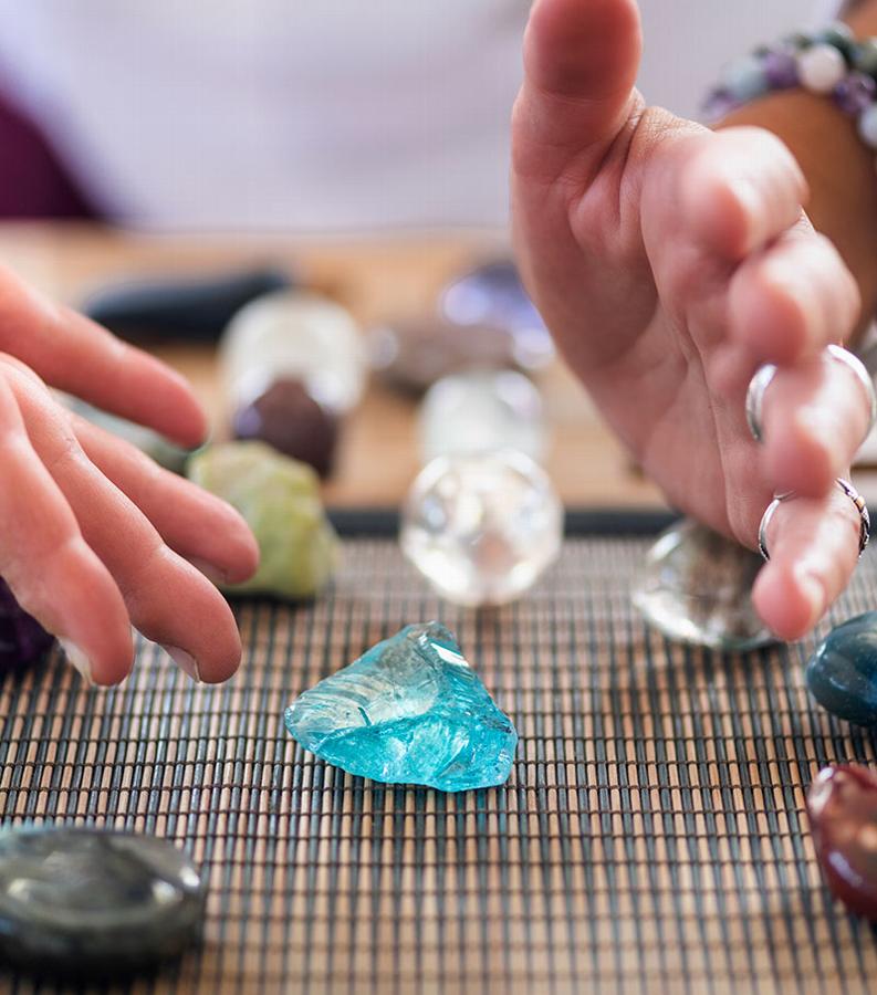Hands reaching towards a blue crystal on a textured surface surrounded by various stones.