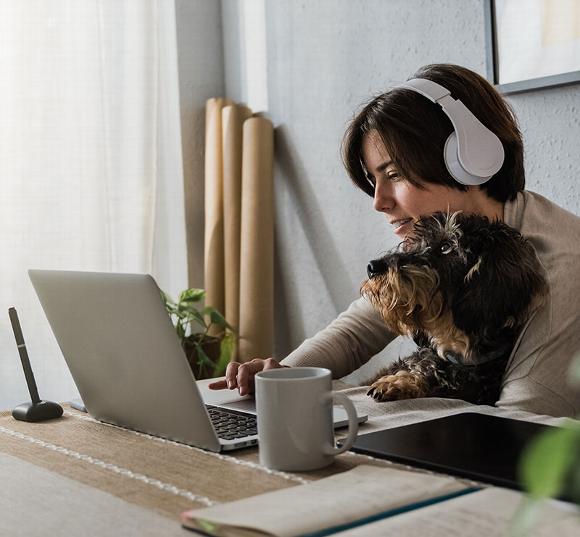 Woman with headphones using a laptop, with a dog sitting beside her on the table.